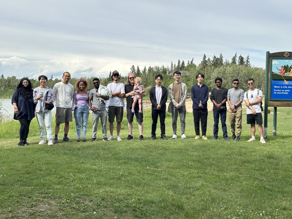 The IDOBE group standing in a line on the grass beside a lake at the 2025 summer BBQ