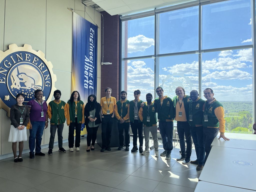 The IDOBE group in green and gold jackets beside the Engineering crest, in front of tall windows overlooking the river valley