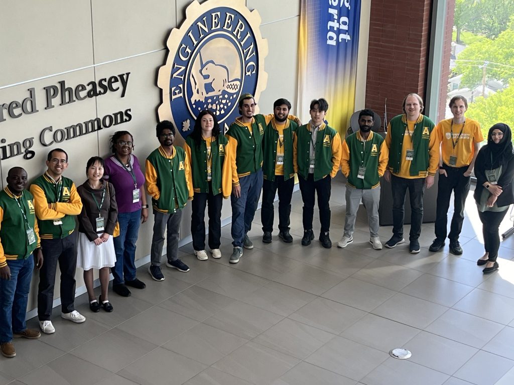 The IDOBE group in green and gold University of Alberta jackets, standing in front of the Faculty of Engineering crest