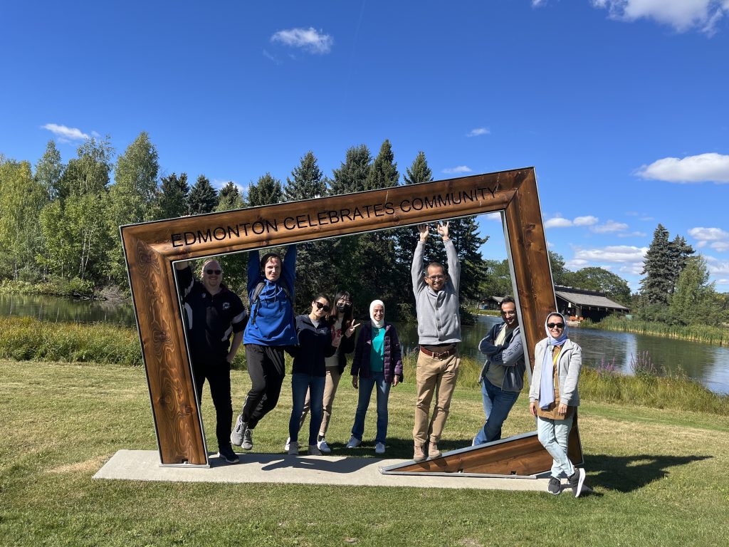 Group members posing inside a large outdoor picture-frame sculpture beside a pond