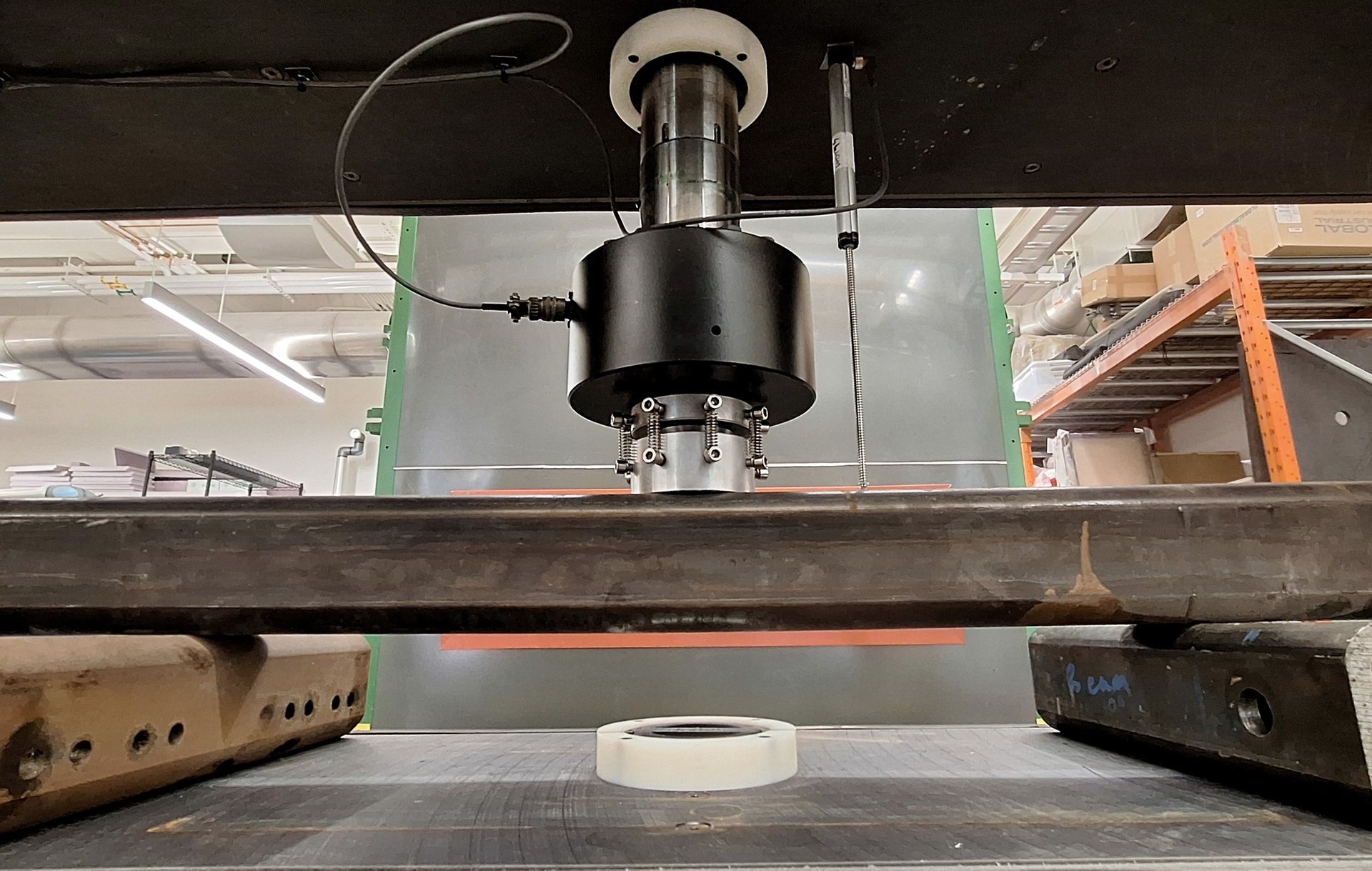 A steel beam tested simply supported in bending, with the load cell and spherical platen bearing down at midspan inside the enclosure.