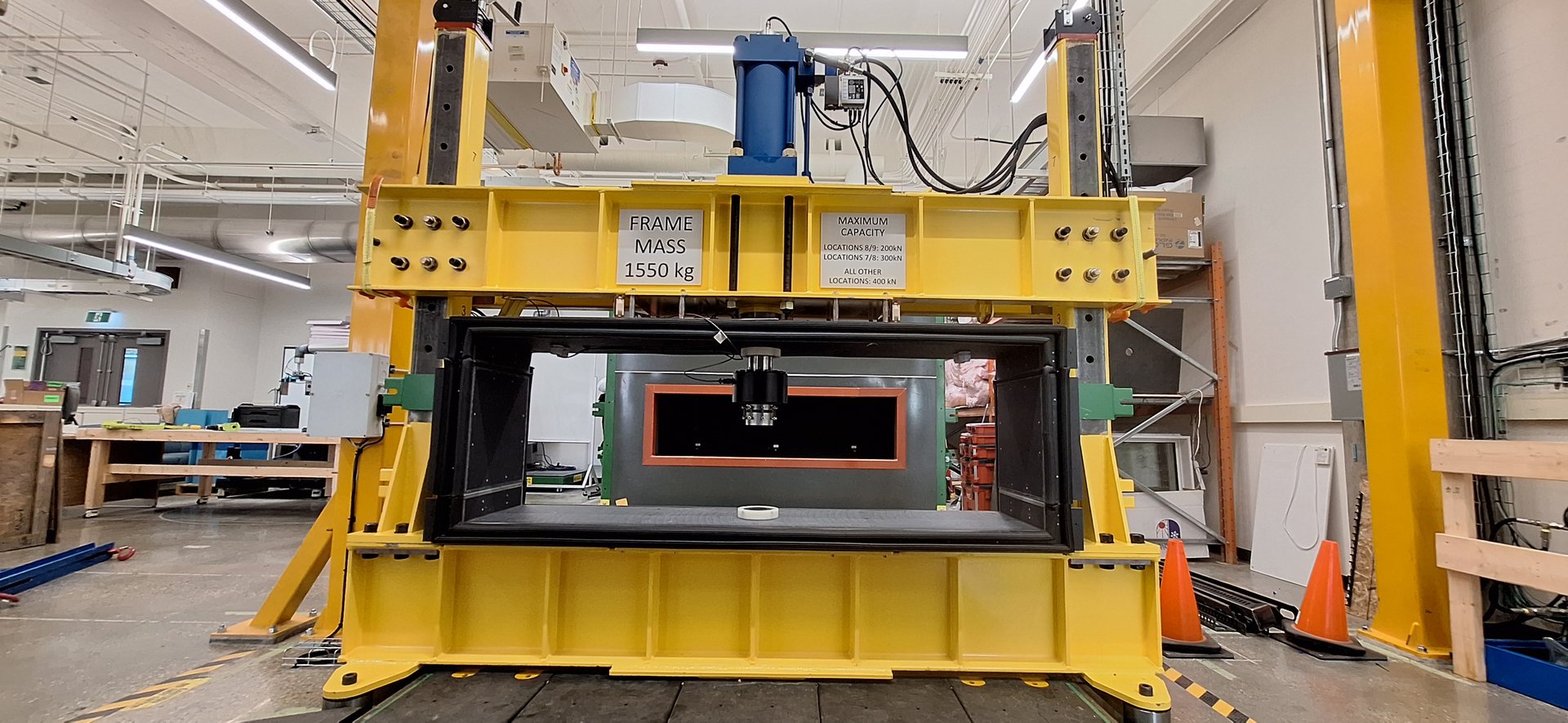 The yellow structural load frame, with its hydraulic cylinder on the crosshead above the sealed test enclosure.